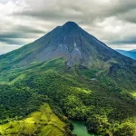 Aerial view of a conical, forested mountain surrounded by lush greenery and a small river, under a cloudy sky.