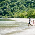Dad and daughter walking along a peaceful beach in Papagayo, Costa Rica, surrounded by lush green jungle.